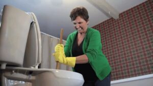 A homeowner in green sweater and yellow rubber gloves, plunging a toilet.