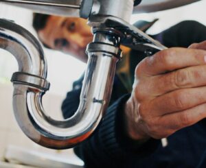 Plumber's hands using wrench to repair silver pipe under a sink in a home.